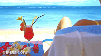 A Woman Sits On A Beach With A Drink On A Table GIF