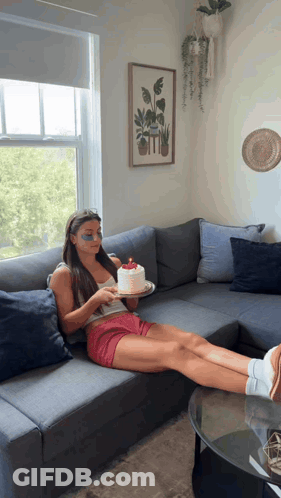 A Woman Sits On A Couch Holding A Cake GIF