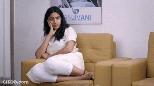 A Woman Sits On A Couch In Front Of A Poster That Says Anjivani GIF