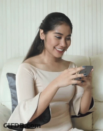 A Woman Sits On A Couch Looking At Her Phone And Smiling GIF