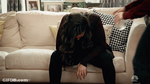 A Woman Sits On A Couch With Her Head On Her Knees And A Nbc Logo On The Bottom Right GIF
