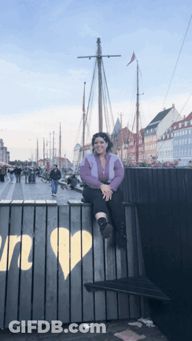 A Woman Sits On A Fence With A Heart Painted On It In Front Of A Boat GIF