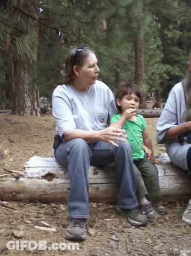 A Woman Sits On A Log Holding A Child Who Is Eating Something GIF