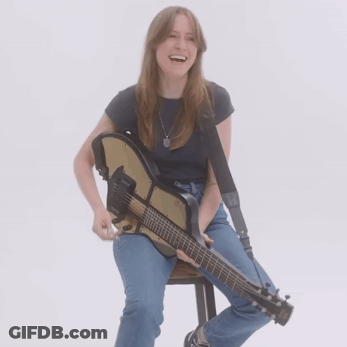 A Woman Sits On A Stool Holding A Guitar And Smiling GIF
