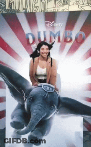 A Woman Sits On A Stuffed Elephant In Front Of A Dumbo Poster GIF