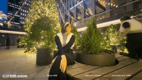 A Woman Sits On A Wooden Bench In Front Of A Christmas Tree With A Watermark That Says Differentwalking GIF