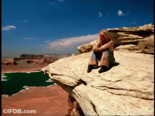 A Woman Sits On The Edge Of A Rocky Cliff Overlooking A Lake GIF