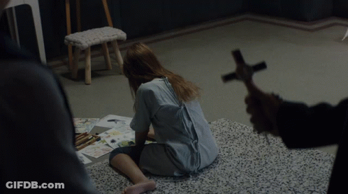 A Woman Sits On The Floor While A Man Holds A Cross In Front Of Her GIF