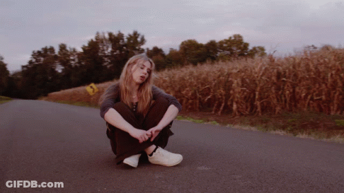 A Woman Sits On The Side Of A Road In Front Of A Cornfield GIF