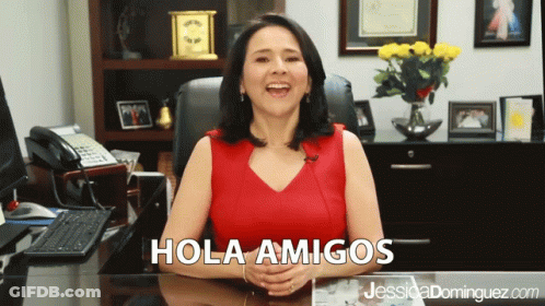 A Woman Sitting At A Desk With Hola Amigos Written On The Top GIF