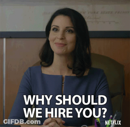 A Woman Sitting At A Desk With The Words " Why Should We Hire You " GIF
