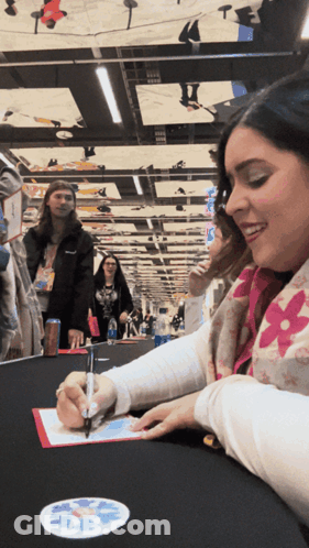 A Woman Sitting At A Table Signing A Piece Of Paper With A Pen GIF