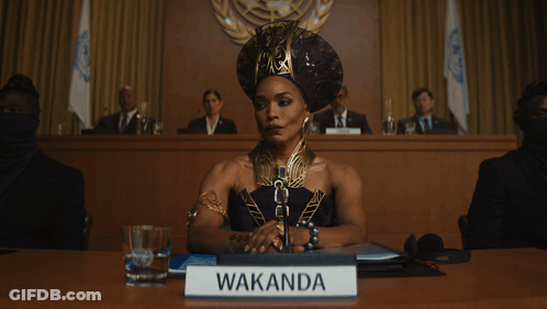 A Woman Sitting At A Table With A Sign That Says Wakanda On It GIF