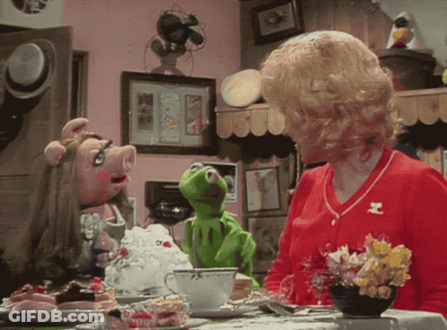 A Woman Sitting At A Table With Muppets Including Kermit GIF