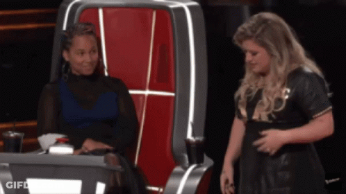 A Woman Sitting In A Chair Talking To Another Woman In A Black Dress GIF