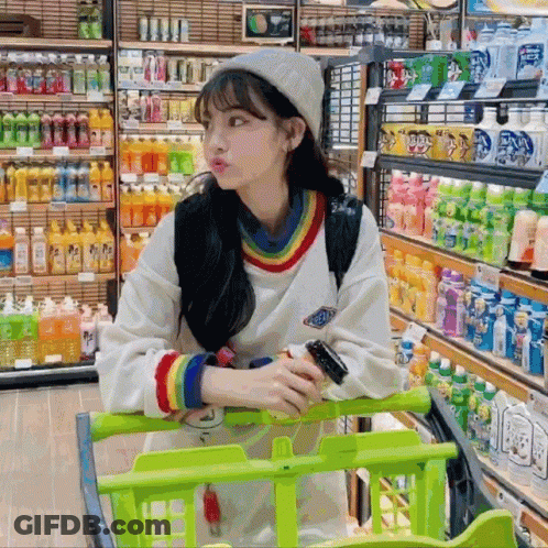A Woman Sitting In A Shopping Cart In A Store With Bottles Of Soda On Shelves GIF