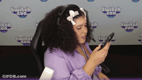 A Woman Sitting In Front Of A Wall That Says ' Twitch Con ' On It GIF