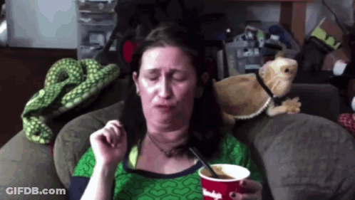 A Woman Sitting On A Couch With A Cup Of Ice Cream And A Spoon In Her Hand GIF