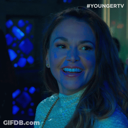 A Woman Smiles And Says " What " In Front Of A Younger TV Logo GIF