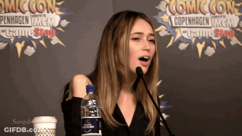 A Woman Speaking Into A Microphone In Front Of A Comic Con Banner GIF