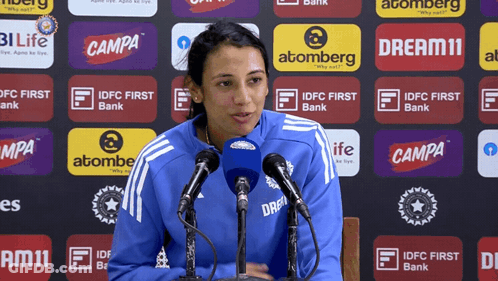 A Woman Speaking Into A Microphone In Front Of A Wall Of Advertisements For Campa And Atomberg GIF
