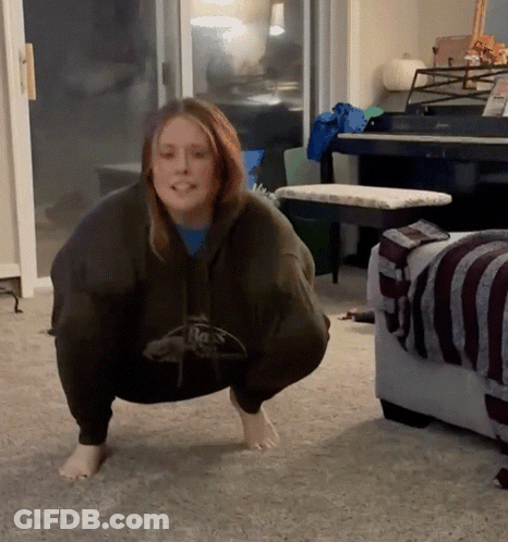 A Woman Squatting Down In A Living Room Wearing A Hoodie That Says Reebok GIF