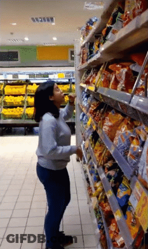 A Woman Standing In A Grocery Store Reaching For A Bag Of Chips GIF