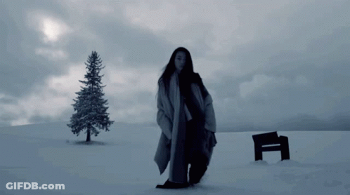A Woman Standing In A Snowy Field With A Bench And A Tree In The Background GIF