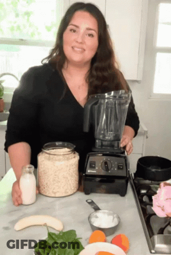 A Woman Standing In Front Of A Blender With A Bottle Of Milk On The Counter GIF