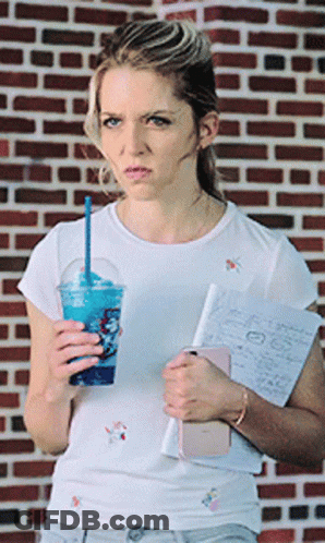 A Woman Standing In Front Of A Brick Wall Holding A Cup Of Blue Liquid GIF