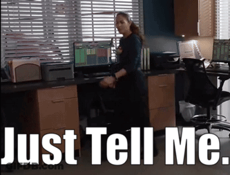A Woman Standing In Front Of A Desk With The Words Just Tell Me Written Above Her GIF