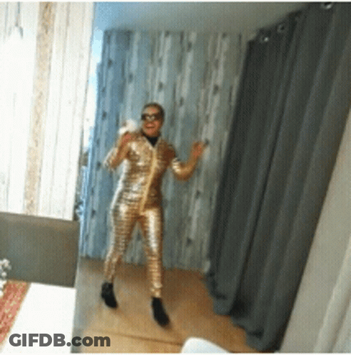 A Woman Standing In Front Of A Mirror Wearing A Gold Jumpsuit GIF
