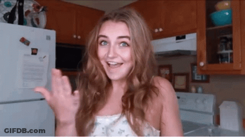 A Woman Standing In Front Of A Refrigerator In A Kitchen Waving Her Hand GIF
