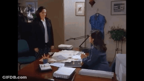 A Woman Standing Next To A Woman Sitting At A Desk In An Office . GIF