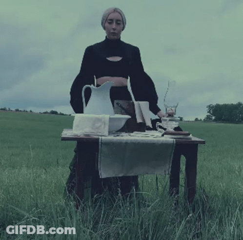 A Woman Stands Behind A Table With A Pitcher And A Lamp GIF