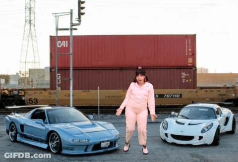 A Woman Stands Between Two Sports Cars In Front Of A Container That Says Cai GIF