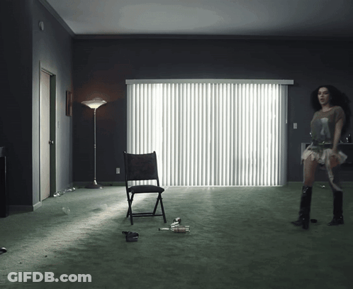 A Woman Stands In A Dark Room With A Chair And A Lamp GIF