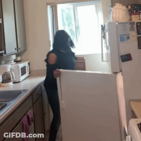 A Woman Stands In A Kitchen Holding A White Refrigerator GIF