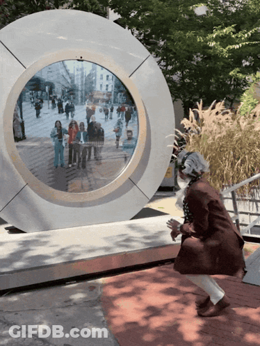 A Woman Stands In Front Of A Circular Sculpture With A Reflection Of People Walking Down A Street GIF