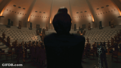 A Woman Stands In Front Of A Large Audience In A Dark Auditorium GIF