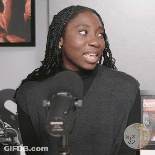 A Woman Stands In Front Of A Microphone With A Creepy Face On The Bottom Right Corner GIF
