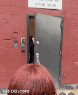 A Woman Stands In Front Of A Stage Door That Is Labeled 212 Ashland Pl GIF