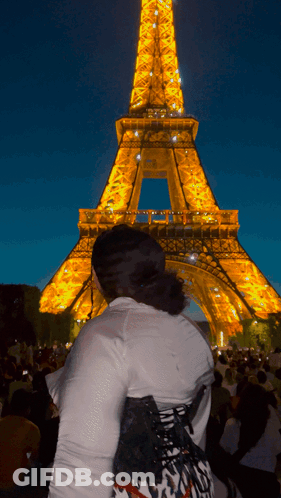 A Woman Stands In Front Of The Eiffel Tower Lit Up At Night GIF