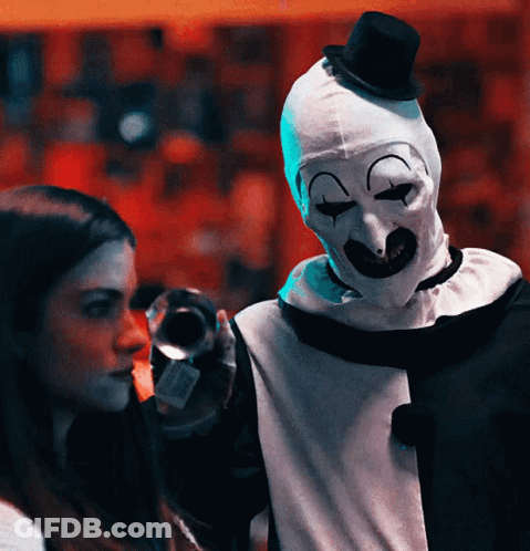 A Woman Stands Next To A Clown Wearing A Mask And Top Hat GIF