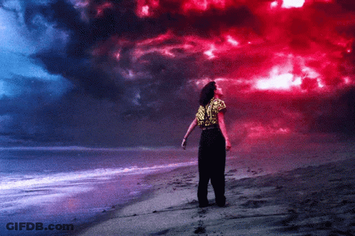 A Woman Stands On A Beach Looking Up At A Red And Blue Sky GIF