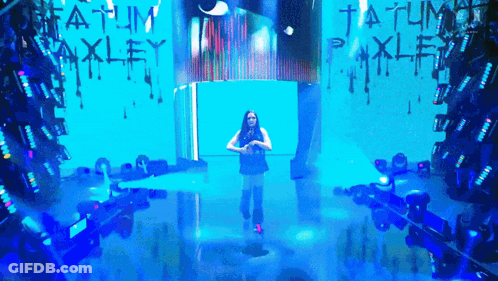 A Woman Stands On A Stage In Front Of A Wall That Says Tatum Oakley GIF