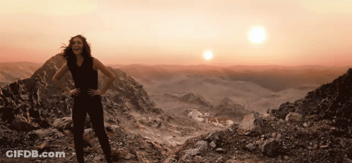 A Woman Stands On Top Of A Rocky Mountain With The Sun Behind Her GIF