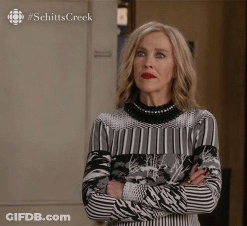 A Woman Stands With Her Arms Crossed In Front Of A Sign That Says Schitts Creek On It GIF