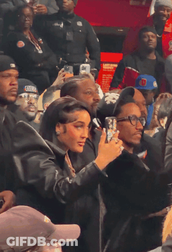 A Woman Taking A Picture Of A Man Wearing A Hat That Says ' Der ' On It GIF