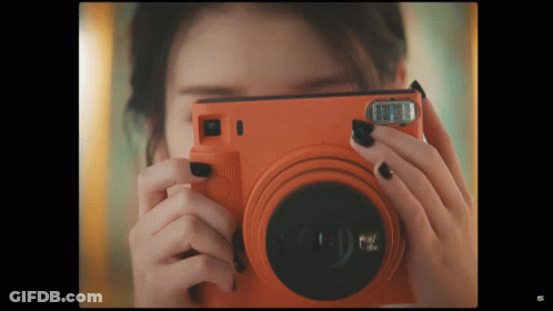 A Woman Taking A Picture With An Orange Camera That Says Fuji GIF